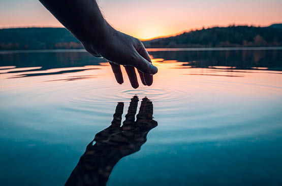 Una mano toca el agua con la punta de los dedos en un atardecer 